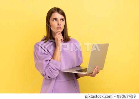 Pensive woman with working on laptop, holding chin, thinking about new project, looking away with thoughtful expression, wearing purple hoodie. Indoor studio shot isolated on yellow background. 99067489