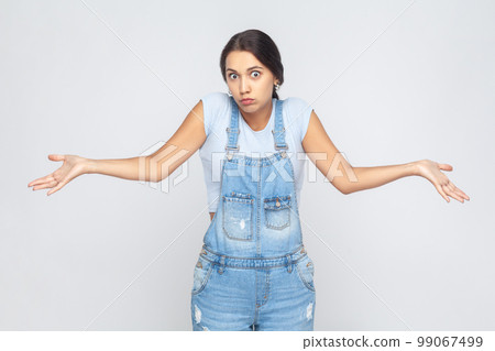 Maybe, don't know. Portrait of unsure woman wearing denim overalls raising hands with confused perplexed puzzled expression, not sure about decision. Indoor studio shot isolated on gray background. Maybe, don't know. Portrait of unsure woman wearing denim overalls raising hands with confused perplexed puzzled expression, not sure about decision. Indoor studio shot isolated on gray background. 99067499