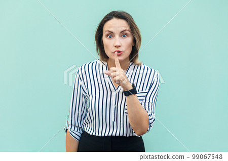 It's a secret. Portrait of serious middle aged woman wearing striped shirt standing with finger near mouth, showing shh gesture. Indoor studio shot isolated on light blue background. 99067548