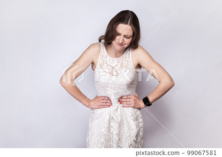 Portrait of sick unhealthy dark haired woman wearing white dress having terrible stomachache, touching her belly, frowning her face. Indoor studio shot isolated on gray background. 99067581