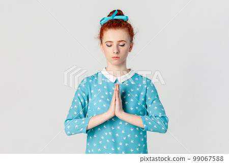 Portrait of calm peaceful attractive ginger woman with bun hairstyle, practicing yoga, keeps eyes closed, meditating, wearing blue dress. Indoor studio shot isolated on gray background. Portrait of calm peaceful attractive ginger woman with bun hairstyle, practicing yoga, keeps eyes closed, meditating, wearing blue dress. Indoor studio shot isolated on gray background. 99067588