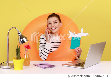Positive woman with rubber ring, holding card with sale inscription, passport and paper plane, talking phone with happy look. Indoor studio studio shot isolated on yellow background. 99067591