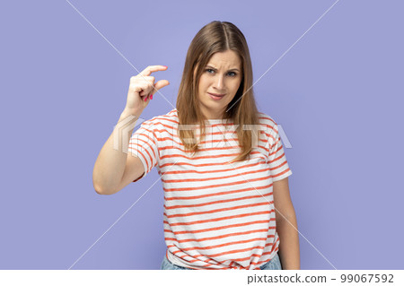 Portrait of beautiful attractive blond woman wearing striped T-shirt showing small gesture with frowning face, being dissatisfied. Indoor studio shot isolated on purple background. 99067592