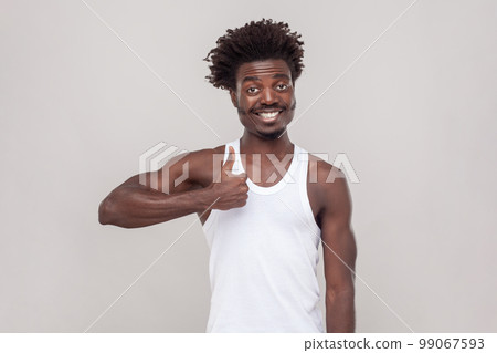 Nice joke. Man with Afro hairstyle showing like gesture, satisfied with something, being in good mood, shows her agreement, wearing white T-shirt. Indoor studio shot isolated on gray background. 99067593