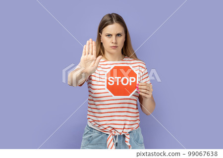 Portrait of blond woman wearing striped T-shirt holding red stop symbol and swing gesture with palm, forbidden sign with hand. Indoor studio shot isolated on purple background. 99067638