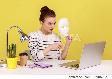Strict woman office worker holding white face mask and pointing finger at laptop screen, blaming partner for hiding identity, duplicity. Indoor studio studio shot isolated on yellow background. 99067640
