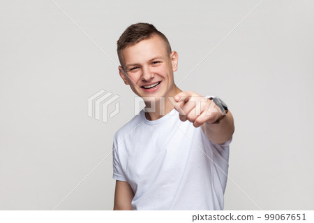Portrait of satisfied smiling positive young teenager boy in T-shirt looking at camera with toothy smile, pointing at camera with finger, choosing you. Indoor studio shot isolated on gray background. Portrait of satisfied smiling positive young teenager boy in T-shirt looking at camera with toothy smile, pointing at camera with finger, choosing you. Indoor studio shot isolated on gray background. 99067651
