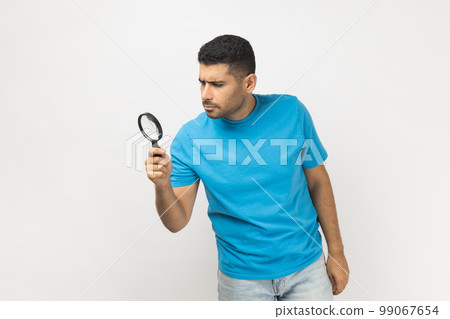 Portrait of unshaven man wearing blue T- shirt standing looking through magnifying glass, finding out something, exploring, inspecting. Indoor studio shot isolated on gray background. 99067654