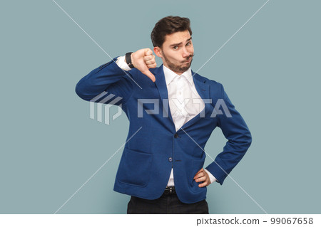 Portrait of dissatisfied attractive man with mustache with frowning face, showing thumb up, negative feedback, wearing white shirt and jacket. Indoor studio shot isolated on light blue background. 99067658