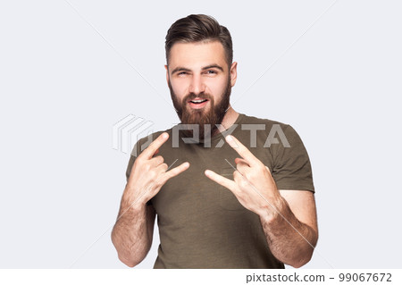 Portrait of naughty excited bearded man wearing dark green T-shirt showing rock and roll gesture with fingers and smiling, attending rock festival. Indoor studio shot isolated on light gray background Portrait of naughty excited bearded man wearing dark green T-shirt showing rock and roll gesture with fingers and smiling, attending rock festival. Indoor studio shot isolated on light gray background 99067672