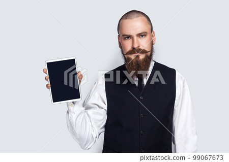 Portrait of serious assertive handsome man with beard and mustache showing tablet with empty display, advertisement area, copy space for promotion. Indoor studio shot isolated on gray background. 99067673