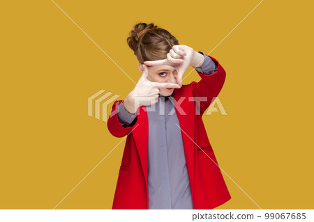 Portrait of concentrated attentive woman with red lips standing looking at camera trough frame from finger, photographing, wearing red jacket. Indoor studio shot isolated on yellow background. Portrait of concentrated attentive woman with red lips standing looking at camera trough frame from finger, photographing, wearing red jacket. Indoor studio shot isolated on yellow background. 99067685