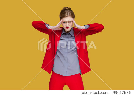Portrait of sad upset unhealthy woman with red lips standing with hands on temples, suffering terrible pain, having headache, wearing red jacket. Indoor studio shot isolated on yellow background. Portrait of sad upset unhealthy woman with red lips standing with hands on temples, suffering terrible pain, having headache, wearing red jacket. Indoor studio shot isolated on yellow background. 99067686
