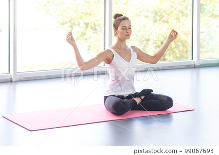 Portrait of relaxed woman wearing sportswear meditating while doing yoga, sitting in lotus pose doing meditation practice, keeps eyes closed. Indoor shot with window on background. 99067693