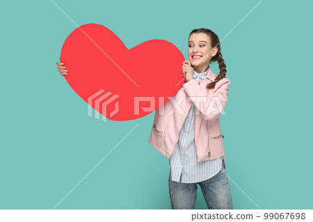 Portrait of extremely happy cheerful teenager girl with braids wearing pink jacket, showing big red heart, looking away with toothy smile. Indoor studio shot isolated on green background. Portrait of extremely happy cheerful teenager girl with braids wearing pink jacket, showing big red heart, looking away with toothy smile. Indoor studio shot isolated on green background. 99067698