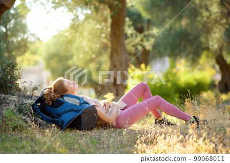 Young woman hiking in countryside. Girl is lying under the tree and having rest and relax. Concepts of adventure, extreme survival, orienteering. Single travel. Backpacking Young woman hiking in countryside. Girl is lying under the tree and having rest and relax. Concepts of adventure, extreme survival, orienteering. Single travel. Backpacking 99068011