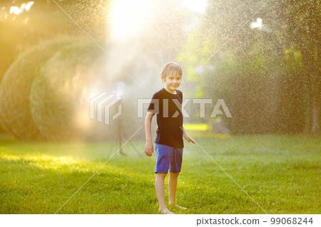 Funny little boy playing with garden sprinkler in sunny city park. Elementary school child laughing, jumping and having fun with spray of water. Summer outdoors activity for kids. 99068244