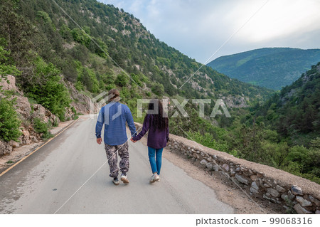 A couple in the mountains rejoice in their journey. They hold hands, stand with their backs to the audience, walk along the road and look at the mountains. A couple in the mountains rejoice in their journey. They hold hands, stand with their backs to the audience, walk along the road and look at the mountains. 99068316