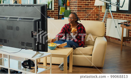 Smiling girl enjoying hamburger and beer bottle, serving fast food takeout order and binge watching tv show. Relaxed woman eating cheeseburger with fries, drinking alcohol at television. 99068813