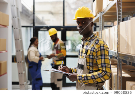 African american man writing on clipboard in industrial warehouse portrait. Post office storehouse package handler wearing helmet using orders checklist and looking at camera 99068826