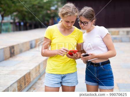 Two young girls with smartphones standing in park 99070625