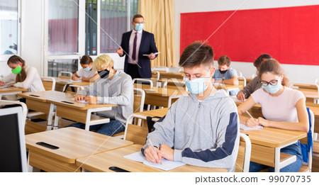 Teenagers in protective masks studying in classroom with teacher Teenagers in protective masks studying in classroom with teacher 99070735