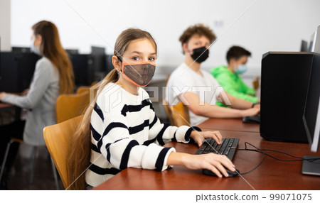 Group of young girls and boys in face masks sitting in computer classroom of library 99071075