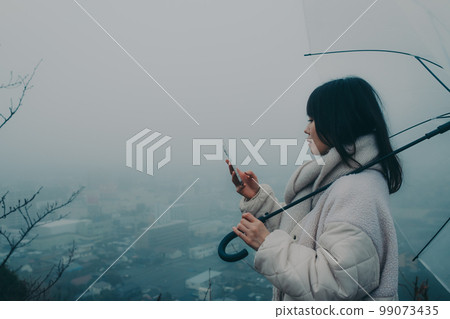 Photo of a woman holding an umbrella in the background of the city and touching her smartphone 99073435
