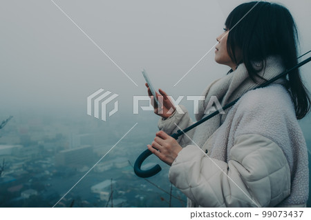 Photo of a woman holding an umbrella in the background of the city and touching her smartphone Photo of a woman holding an umbrella in the background of the city and touching her smartphone 99073437