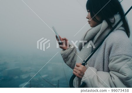 照片中一位女士在城市背景中打著雨傘並觸摸她的智能手機 照片中一位女士在城市背景中打著雨傘並觸摸她的智能手機 99073438