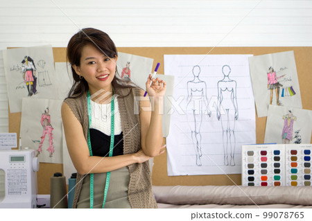 Young fashion designer holding magic pen with a smile. Pattern for cloth and color sample catalog are on cork board. Asian woman working in home base cloth design business. Young fashion designer holding magic pen with a smile. Pattern for cloth and color sample catalog are on cork board. Asian woman working in home base cloth design business. 99078765