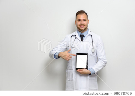 Young caucasian doctor with short hair, mustache and beard dressed  in white gown and stethoscope pointing finger at tablet computer monitor. Stand in front of white wall. 99078769