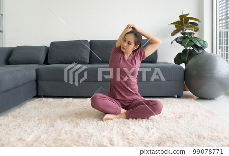 Asian housewife stretching while sitting during home workout on carpet in living room. 99078771