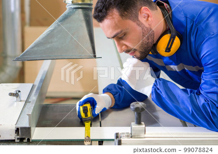 Young technician in blue mechanic jumpsuit, protective glove and ear muff use tape measure to measure the size of wood veneer on a large sliding table saw machine. Atmosphere in furniture factory. Young technician in blue mechanic jumpsuit, protective glove and ear muff use tape measure to measure the size of wood veneer on a large sliding table saw machine. Atmosphere in furniture factory. 99078824
