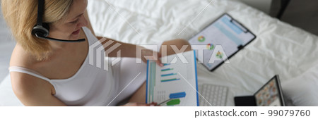 Woman in headphones holds charts sits on bed and holds online conference 99079760