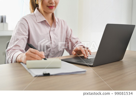 Smiling asian woman worker using laptop computer and making notes on notebook at her workstation Smiling asian woman worker using laptop computer and making notes on notebook at her workstation 99081534