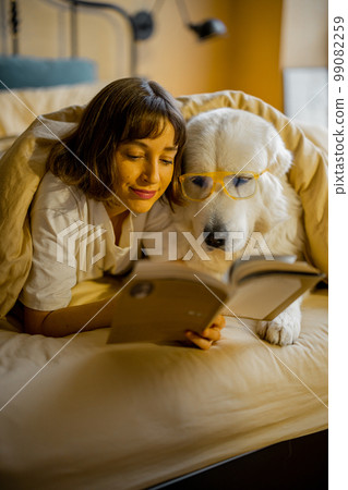 Woman with her cute dog reading book in bed Woman with her cute dog reading book in bed 99082259
