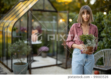 Woman with flowerpot in garden with greenhouse behind 99083526