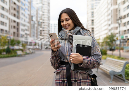 A business woman on the way to work with a laptop in her hands looks at the phone screen with a 99085774