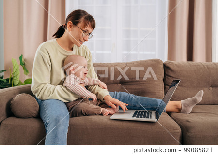 Young mom in glasses using laptop while sitting on couch with her baby at home 99085821