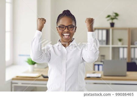 Happy smiling dark-skinned beautiful girl with glasses celebrates victory by clenching her fists and raising her hands up. 99086465