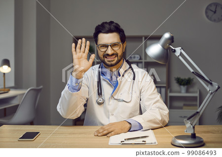 Portrait of cheerful male doctor sitting at table with and waving his hand in greeting Portrait of cheerful male doctor sitting at table with and waving his hand in greeting 99086493