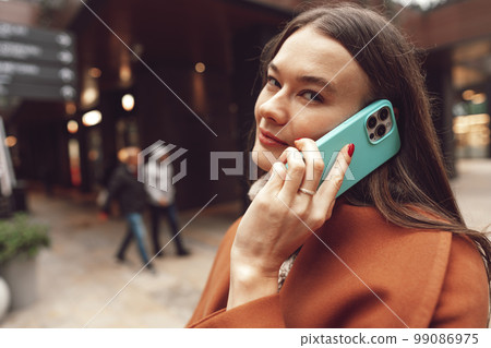Cute brunette woman in brown coat talking on the phone in the city 99086975