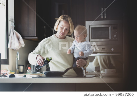 Happy mother and little infant baby boy together making pancakes for breakfast in domestic kitchen. Family, lifestyle, domestic life, food, healthy eating and people concept 99087200