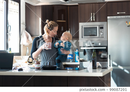 Happy mother and little infant baby boy together making pancakes for breakfast in domestic kitchen. Family, lifestyle, domestic life, food, healthy eating and people concept 99087201