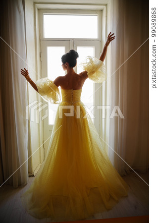A woman's silhouette in a golden luxurious dress against the background of a window holds a curtain with her hands. Elegant lady in a yellow long silk dress with bare back, back view. A woman's silhouette in a golden luxurious dress against the background of a window holds a curtain with her hands. Elegant lady in a yellow long silk dress with bare back, back view. 99089868