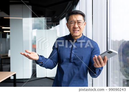 Disappointed man inside office standing near window, portrait of upset asian man inside office with phone, business man looking at camera and spreading hands. Disappointed man inside office standing near window, portrait of upset asian man inside office with phone, business man looking at camera and spreading hands. 99090243