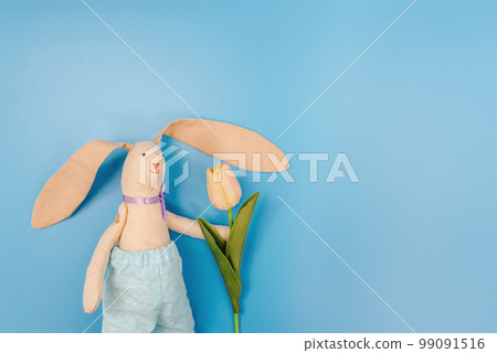 Greeting card Toy bunny with long ears and in pants holding pink tulip on blue background. Concept of spring and Easter. Copy Space for text. Greeting card Toy bunny with long ears and in pants holding pink tulip on blue background. Concept of spring and Easter. Copy Space for text. 99091516
