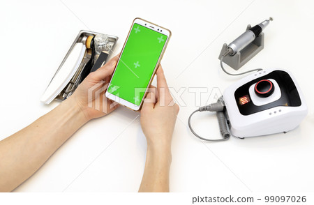 Nail stylist hands hold gadget with green chroma key screen, sitting at workstation in nail beauty treatment salon. Professional tools are on white table. Studying manicurist. Closeup, horizontal 99097026
