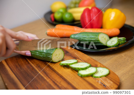 female hands slicing a cucumber on a wooden cutting board 99097301